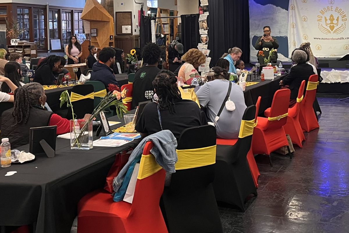 Group of people seated at long banquet tables in a hall, listening to a speaker on a stage with a banner reading 'No More Bound' in the background, flowers on tables and red chairs with yellow sashes.