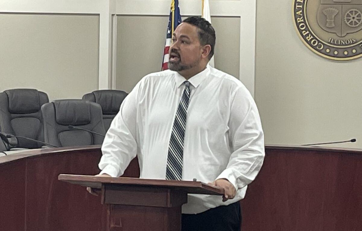 Man in a white shirt and striped tie speaks at a wooden podium in a council chamber, with the US flag and a state seal behind him.