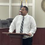 Man in a white shirt and striped tie speaks at a wooden podium in a council chamber, with the US flag and a state seal behind him.