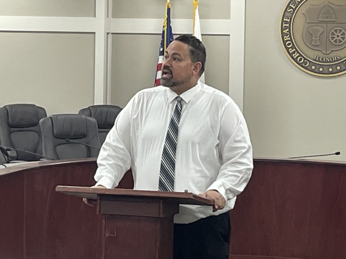 Man in a white shirt and striped tie speaks at a wooden podium in a council chamber, with the US flag and a state seal behind him.