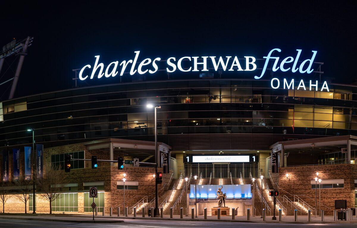 Night exterior of Charles Schwab Field Omaha with illuminated sign atop the stadium and lit entry stairs below, viewed from the street at dusk.
