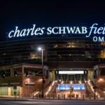 Night exterior of Charles Schwab Field Omaha with illuminated sign atop the stadium and lit entry stairs below, viewed from the street at dusk.