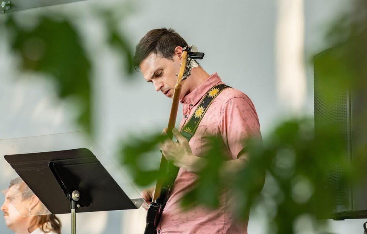 Guitarist in a pink shirt playing an electric guitar on stage, with a music stand in front.