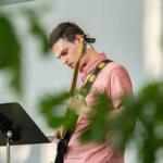 Guitarist in a pink shirt playing an electric guitar on stage, with a music stand in front.