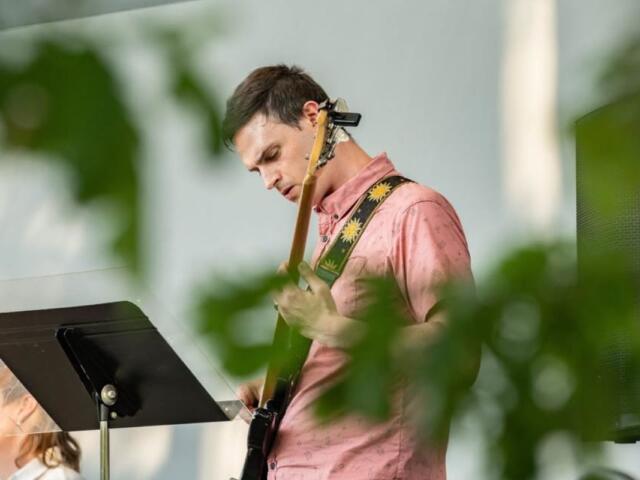 Guitarist in a pink shirt playing an electric guitar on stage, with a music stand in front.