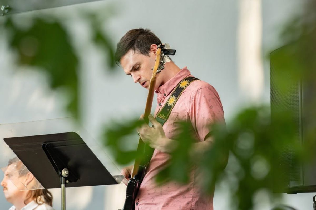 Guitarist in a pink shirt playing an electric guitar on stage, with a music stand in front.