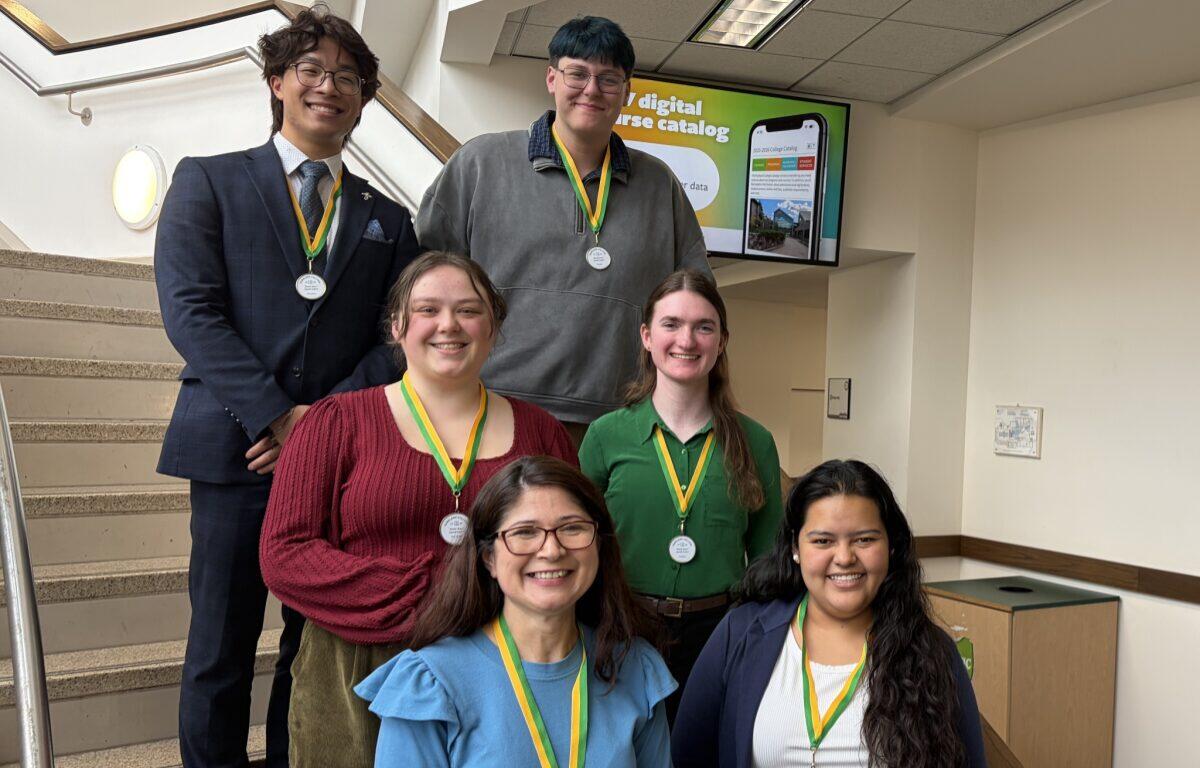 Six young adults stand on a stairwell posing for a group photo, each wearing a medal on a green-and-yellow ribbon after a ceremony or competition.