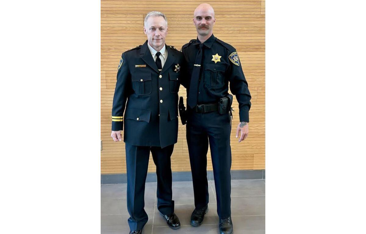 Two police officers in dark dress uniforms stand side by side indoors against a wooden wall.