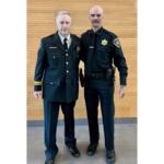Two police officers in dark dress uniforms stand side by side indoors against a wooden wall.