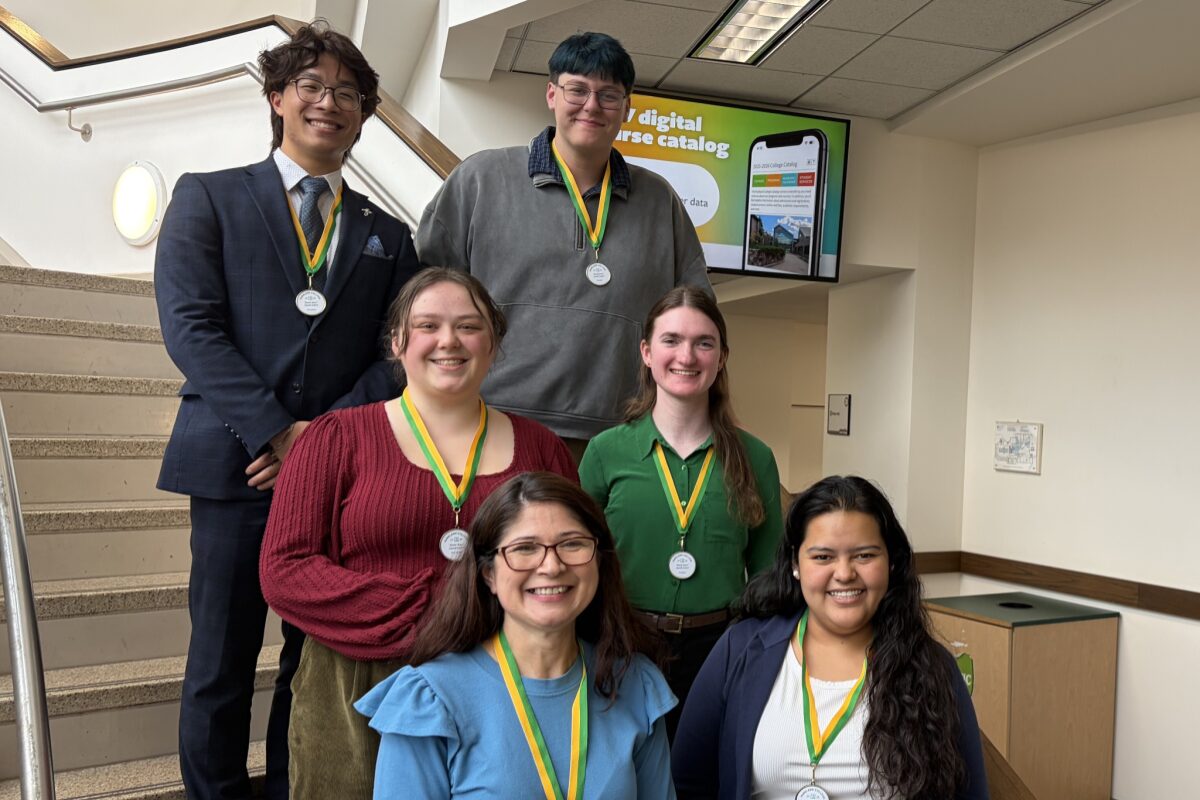 Six young adults stand on a stairwell posing for a group photo, each wearing a medal on a green-and-yellow ribbon after a ceremony or competition.