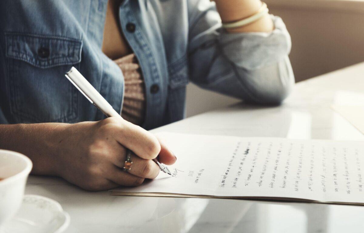 Person signs a document with a pen on a white table, denim shirt sleeve visible and a coffee cup nearby.