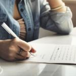 Person signs a document with a pen on a white table, denim shirt sleeve visible and a coffee cup nearby.
