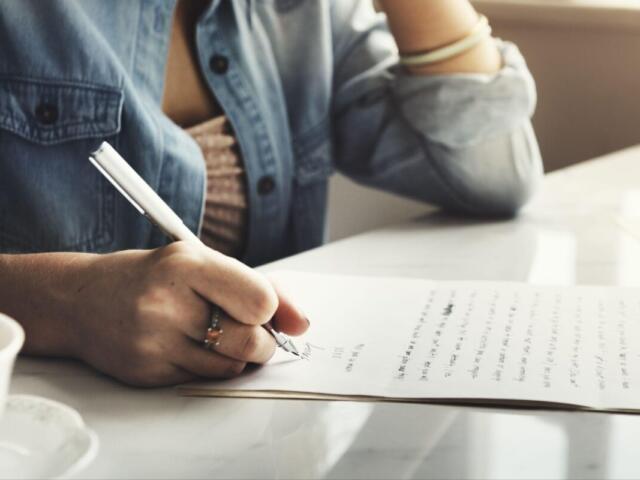 Person signs a document with a pen on a white table, denim shirt sleeve visible and a coffee cup nearby.
