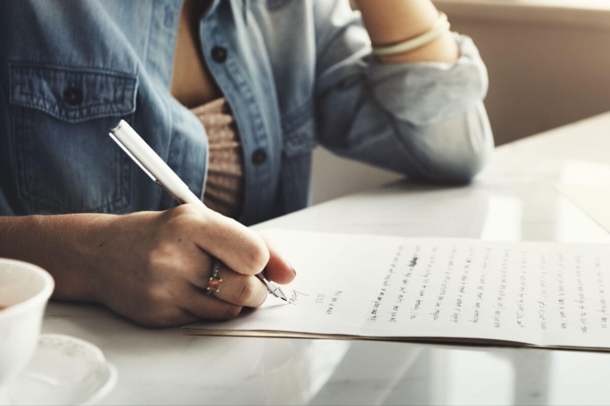 Person signs a document with a pen on a white table, denim shirt sleeve visible and a coffee cup nearby.