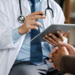 Doctor in a white coat with stethoscope showing a tablet to a patient in a clinic.