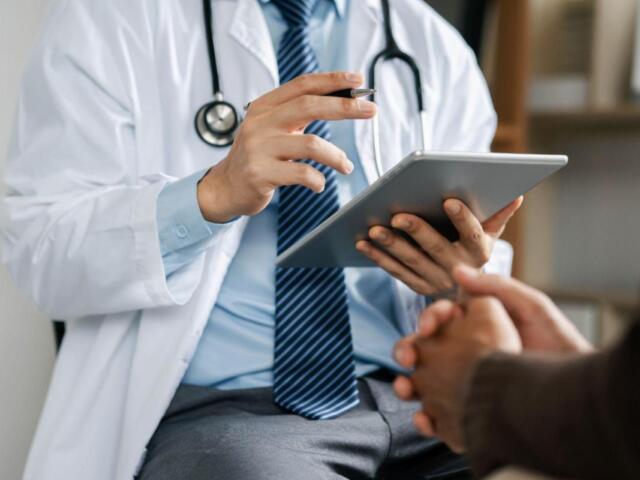 Doctor in a white coat with stethoscope showing a tablet to a patient in a clinic.