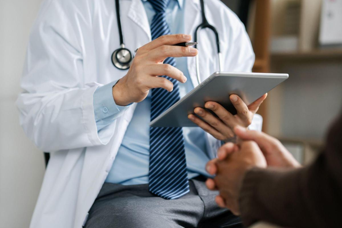 Doctor in a white coat with stethoscope showing a tablet to a patient in a clinic.