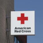 White square sign with a red cross logo and the words 'American Red Cross' mounted to a brick building against a blue sky.