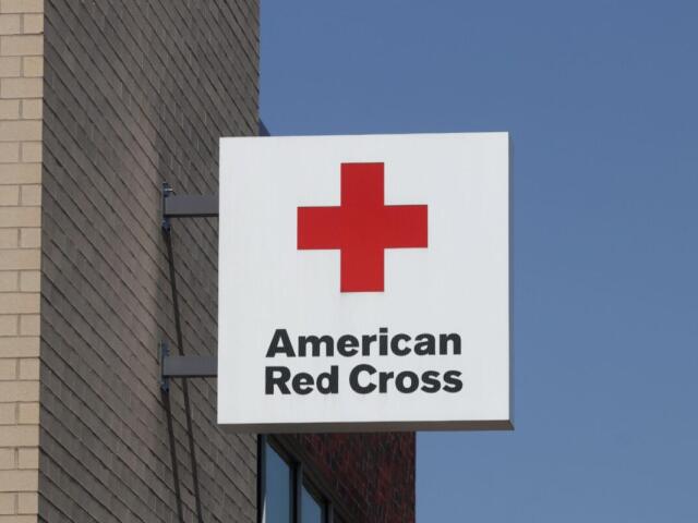 White square sign with a red cross logo and the words 'American Red Cross' mounted to a brick building against a blue sky.