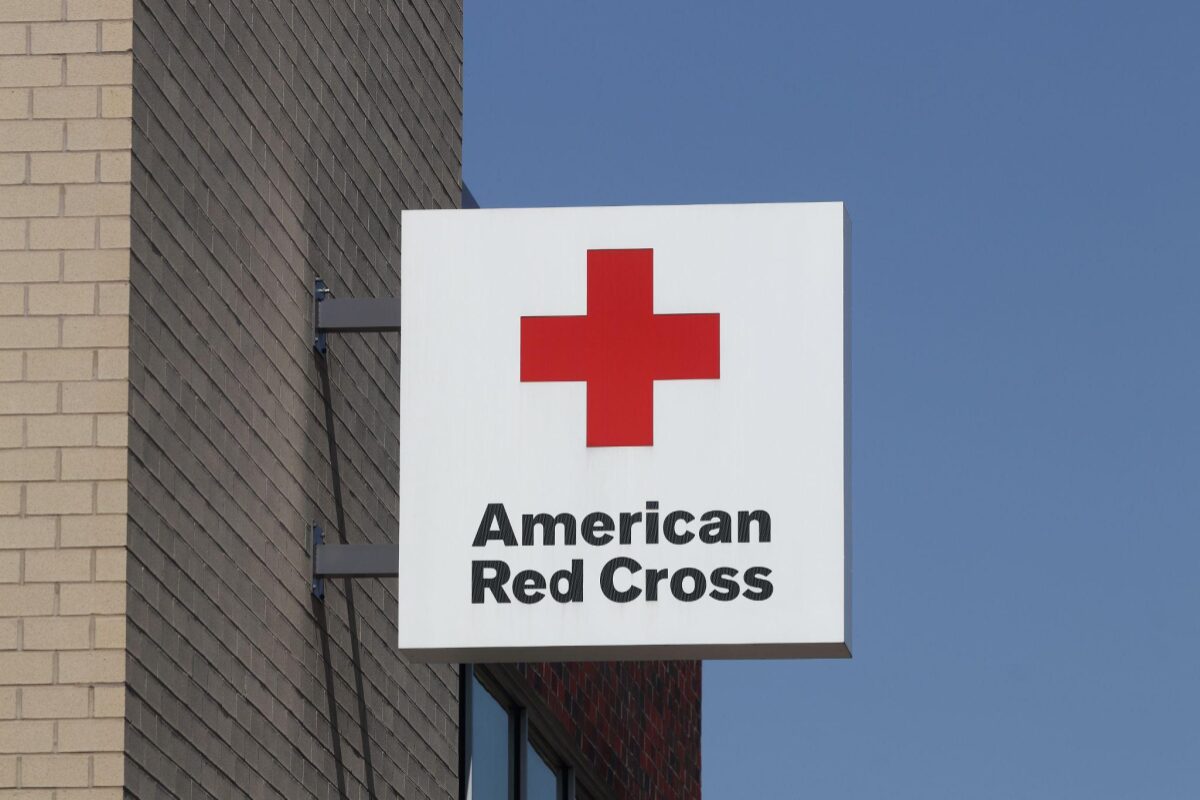 White square sign with a red cross logo and the words 'American Red Cross' mounted to a brick building against a blue sky.