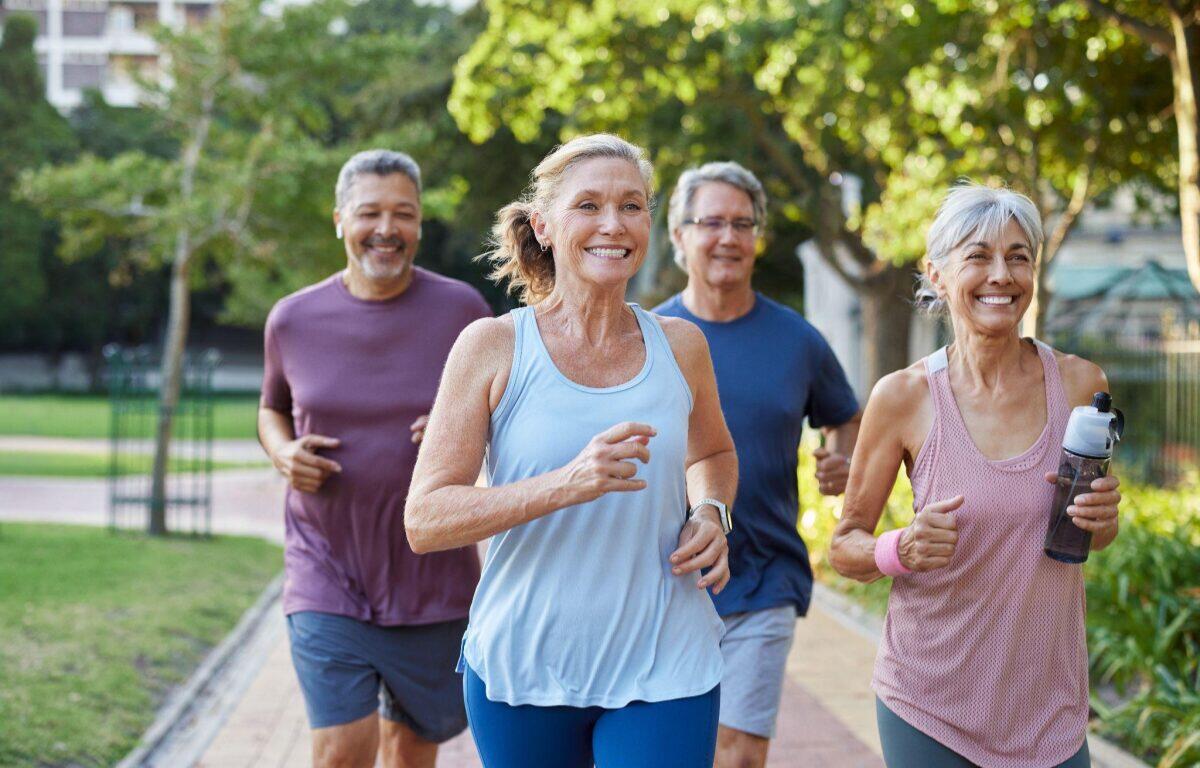 Four adults jogging together on a sunlit park path, smiling as they run.