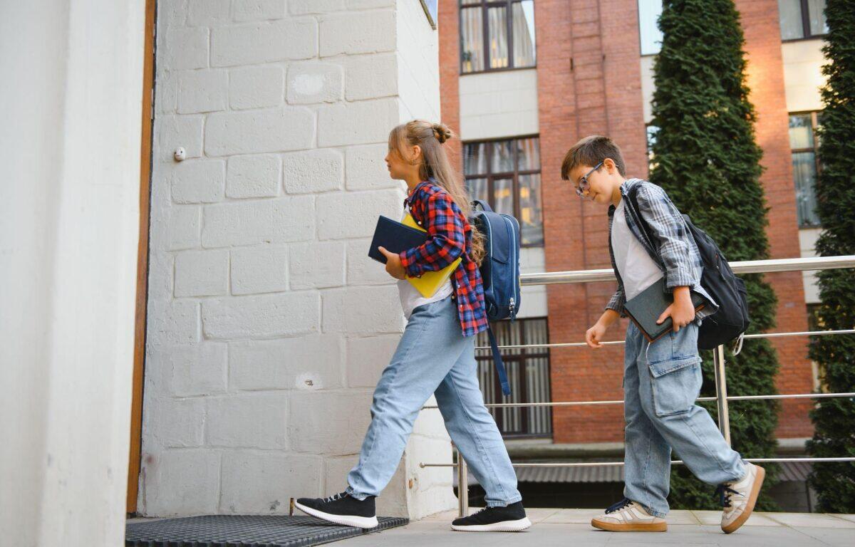 Two students with backpacks walk up a ramp outside a brick building, each carrying books and notebooks.