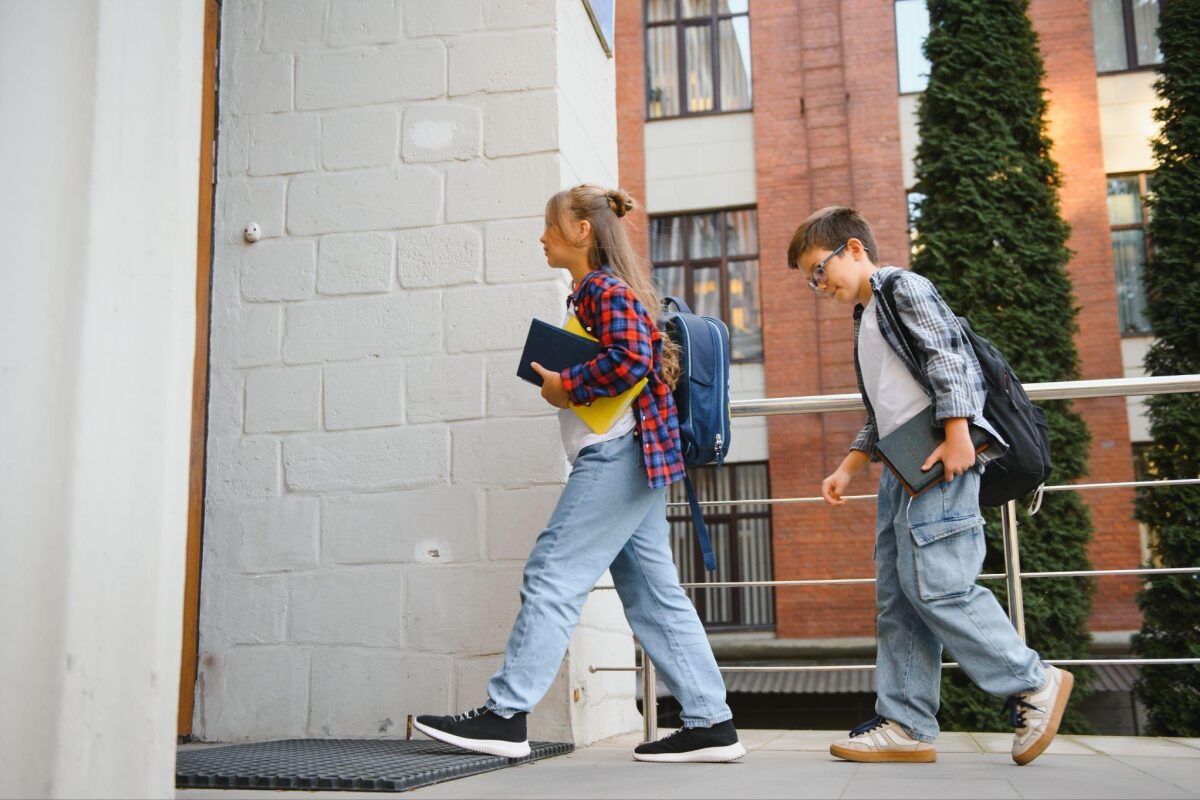 Two students with backpacks walk up a ramp outside a brick building, each carrying books and notebooks.