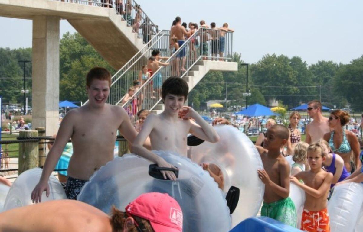 Group of kids and adults in inflatable inner tubes at a crowded outdoor water park, with a tall viewing platform in the background.