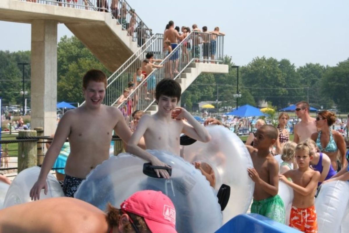 Group of kids and adults in inflatable inner tubes at a crowded outdoor water park, with a tall viewing platform in the background.