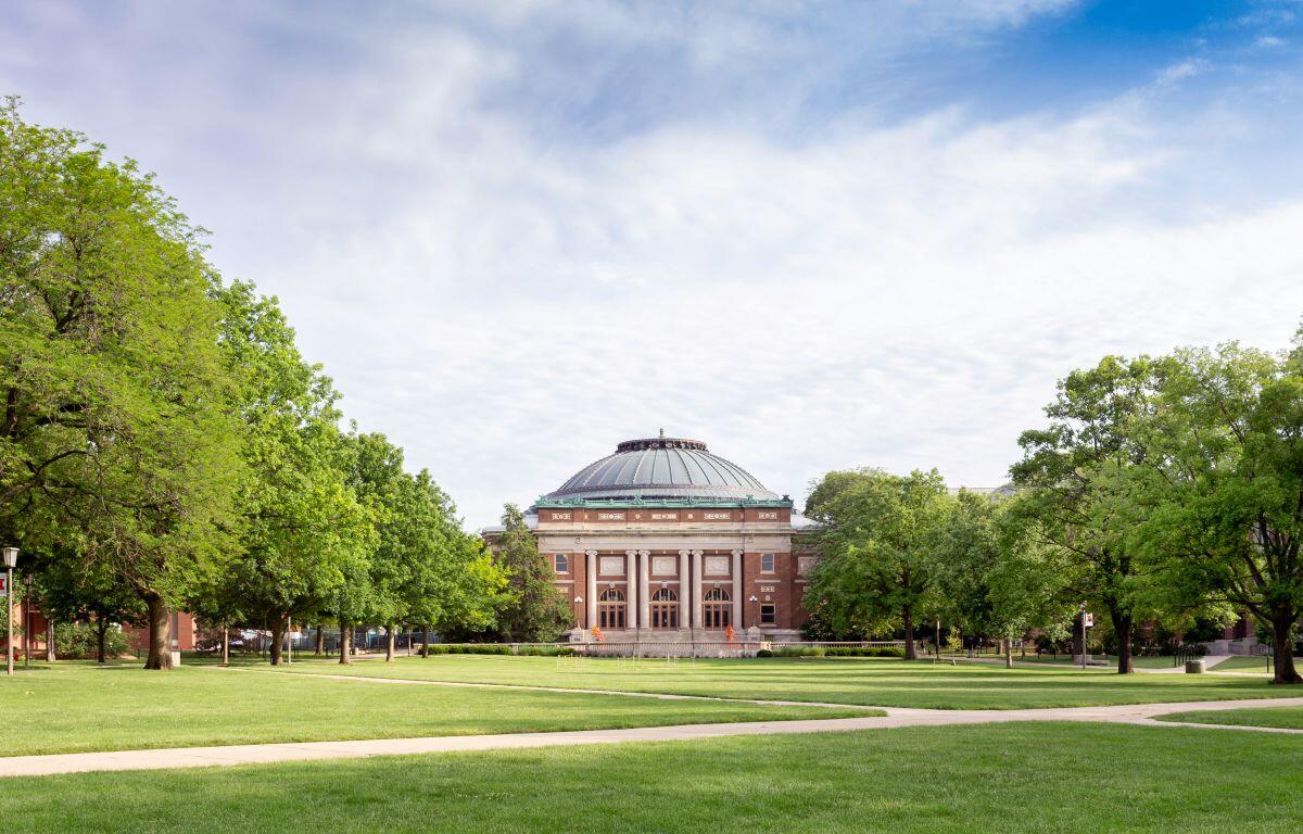 Campus park with a domed brick building centered in a grassy quad, flanked by trees and paved paths.