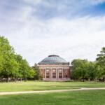 Campus park with a domed brick building centered in a grassy quad, flanked by trees and paved paths.