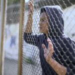 Person in a dark hoodie stands behind a chain-link fence, looking to the side, with hands on the fence.