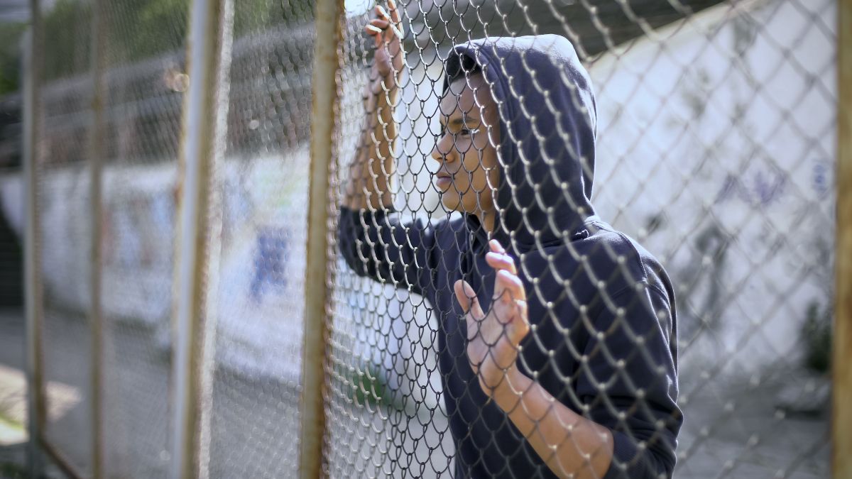 Person in a dark hoodie stands behind a chain-link fence, looking to the side, with hands on the fence.