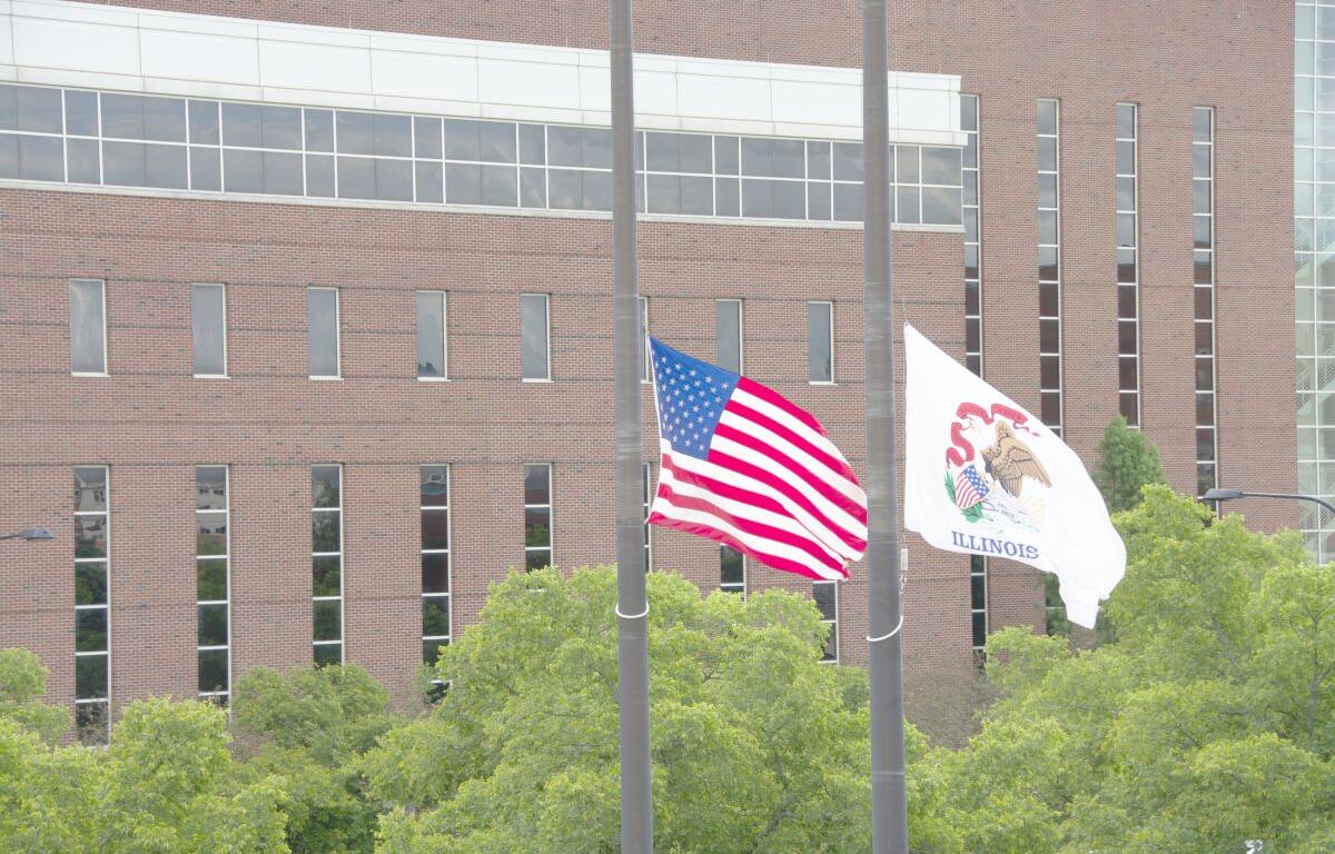 American flag and Illinois state flag flying on flagpoles in front of a brick building with green trees nearby