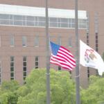 American flag and Illinois state flag flying on flagpoles in front of a brick building with green trees nearby