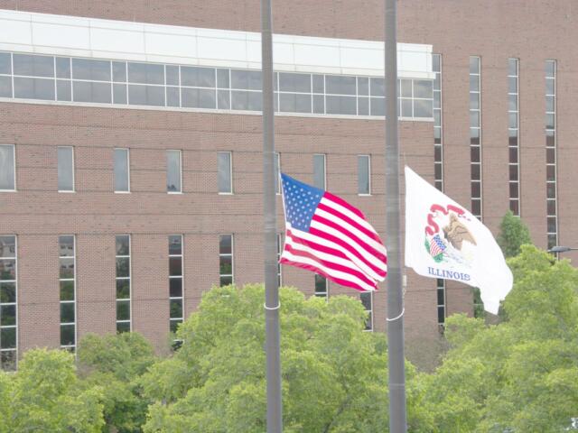 American flag and Illinois state flag flying on flagpoles in front of a brick building with green trees nearby