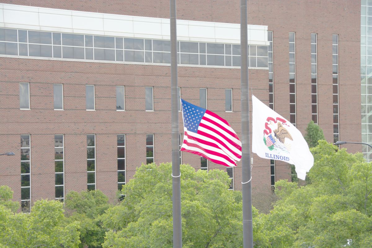 American flag and Illinois state flag flying on flagpoles in front of a brick building with green trees nearby