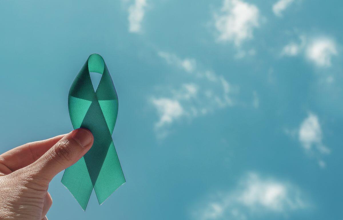 Teal awareness ribbon held between a finger and thumb against a bright blue sky.