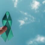 Teal awareness ribbon held between a finger and thumb against a bright blue sky.
