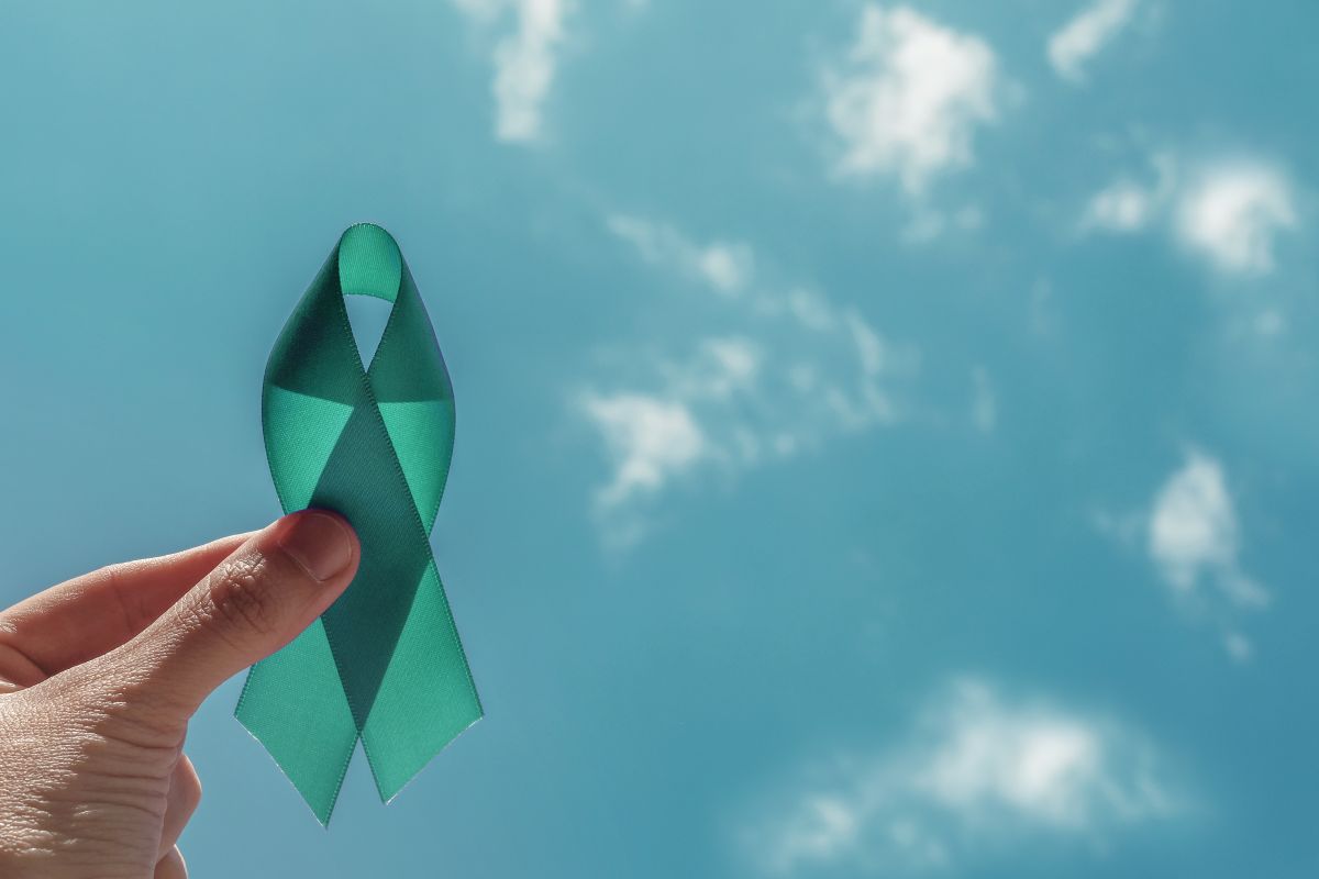 Teal awareness ribbon held between a finger and thumb against a bright blue sky.