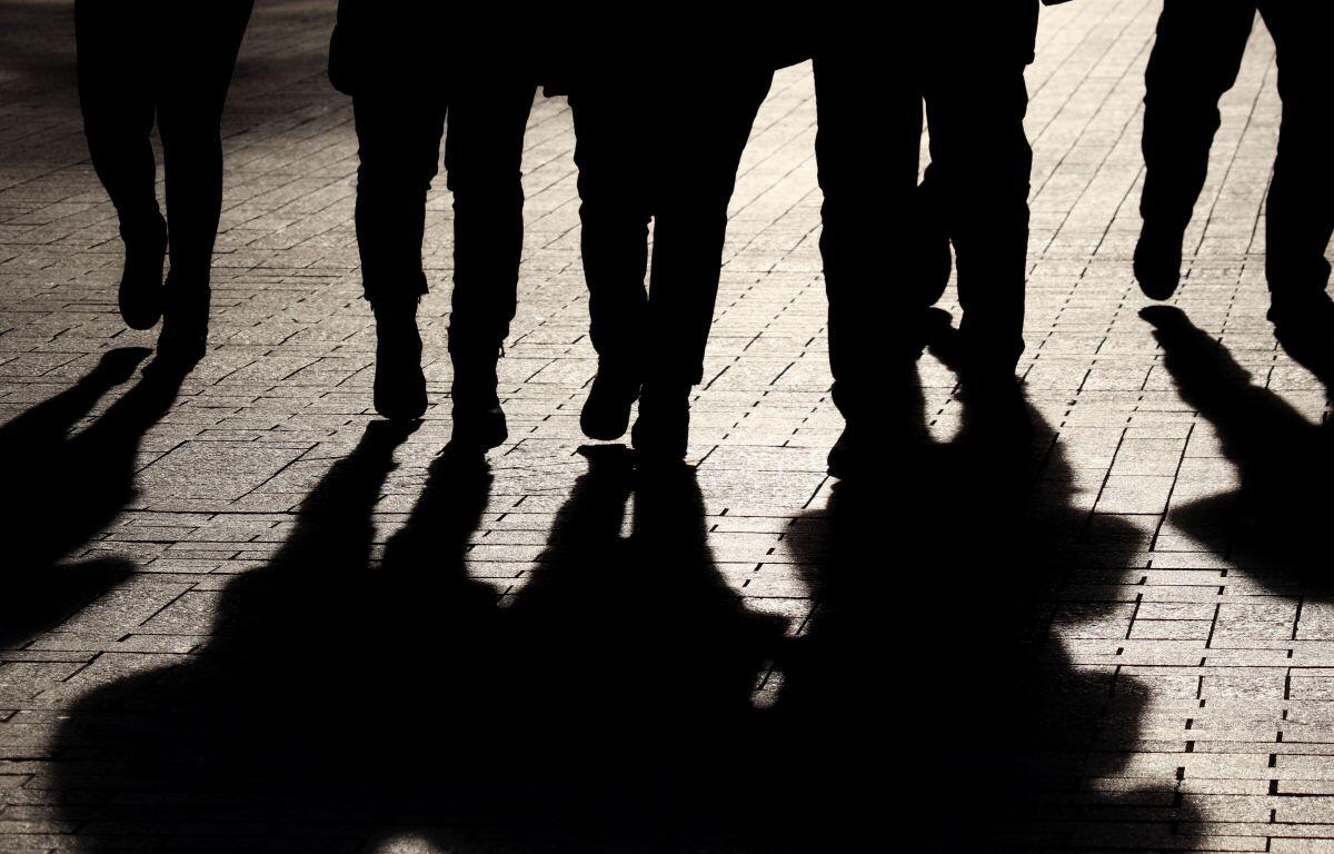 Silhouettes of pedestrians walking on a sunlit brick-paved sidewalk, with long shadows stretching ahead on the ground.