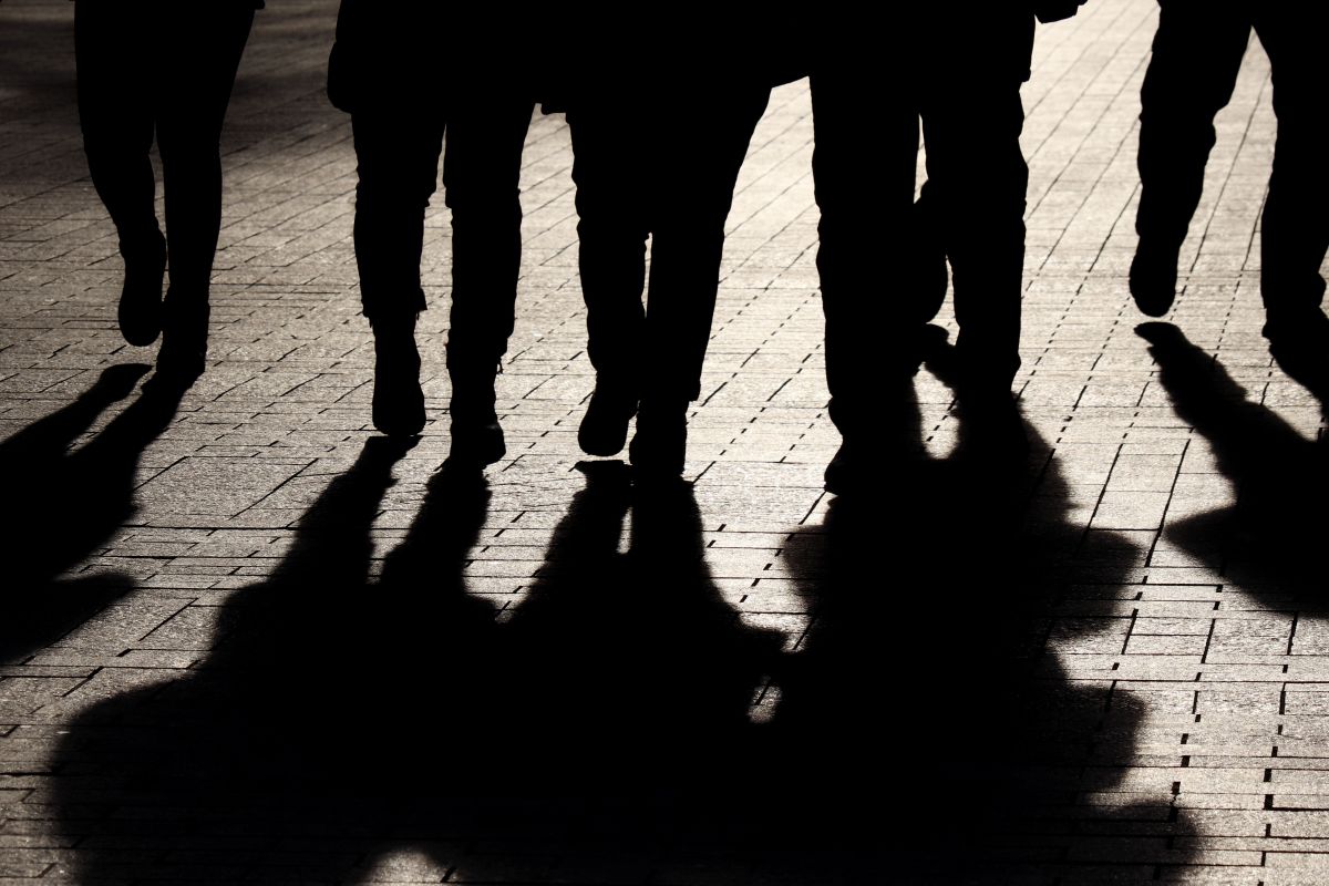 Silhouettes of pedestrians walking on a sunlit brick-paved sidewalk, with long shadows stretching ahead on the ground.