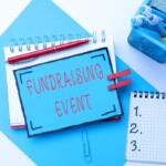 Blue sign reading 'Fundraising Event' placed on a desk with notebooks and office supplies.