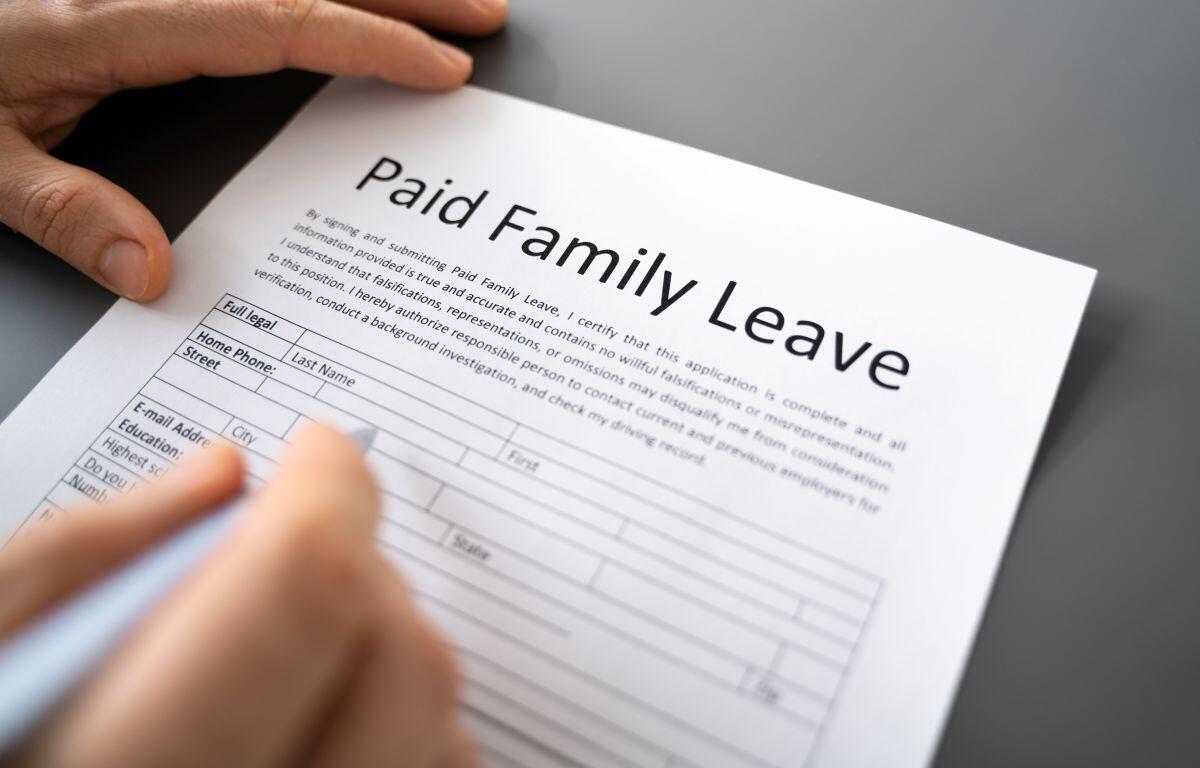 Close-up of a hand filling out a Paid Family Leave form on a gray desk surface, with a blue pen poised over the document.