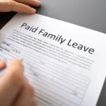 Close-up of a hand filling out a Paid Family Leave form on a gray desk surface, with a blue pen poised over the document.