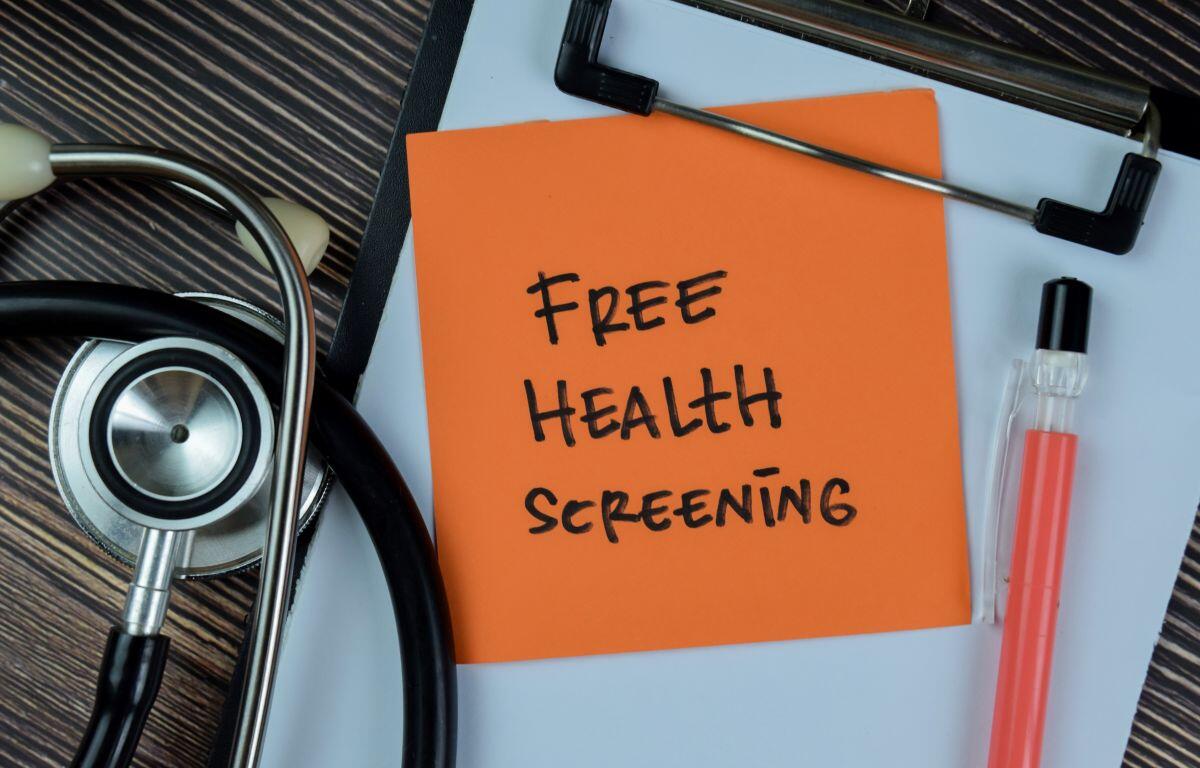 Orange note on a clipboard reads 'Free Health Screening' beside a stethoscope and a pink liquid sample tube nearby.