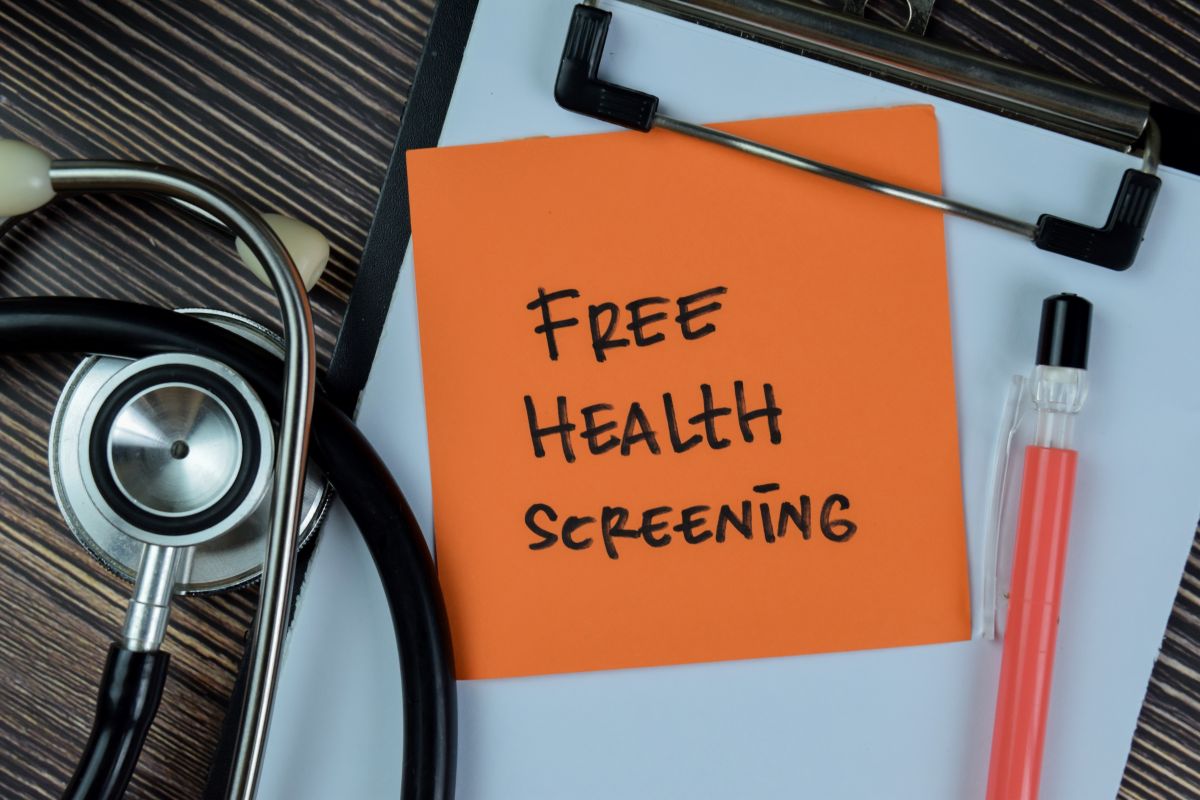 Orange note on a clipboard reads 'Free Health Screening' beside a stethoscope and a pink liquid sample tube nearby.