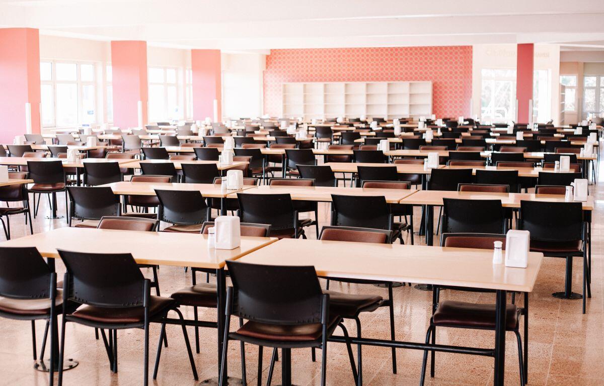 A large cafeteria or dining hall with rows of light wooden tables and black chairs, condiments on each table, bright natural light from windows.
