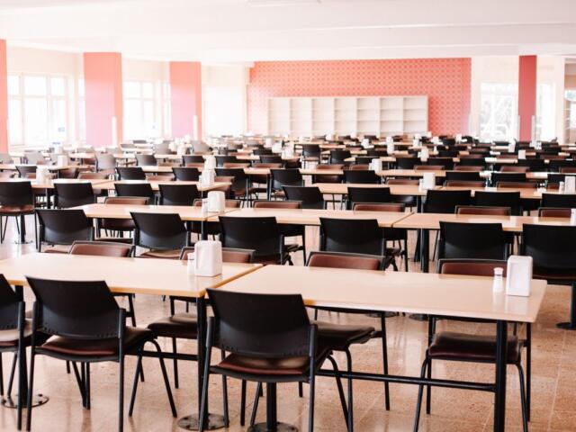 A large cafeteria or dining hall with rows of light wooden tables and black chairs, condiments on each table, bright natural light from windows.