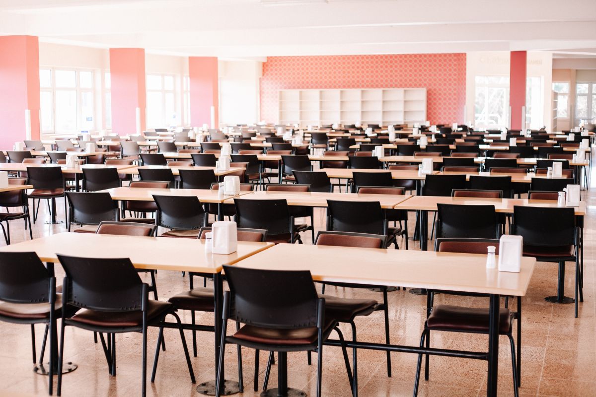 A large cafeteria or dining hall with rows of light wooden tables and black chairs, condiments on each table, bright natural light from windows.